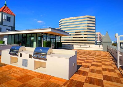 A modern rooftop terrace featuring a built-in grilling area on a sunny day, with a view of city buildings under a clear blue sky.