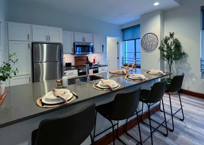 Modern kitchen with stainless steel appliances, gray cabinets, and a central island with bar stools set for dining. there are decorative plants and natural light filtering through large windows.