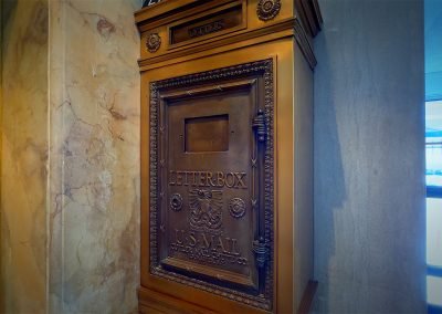 Antique bronze u.s. mail letterbox with intricate designs, mounted on a wooden panel beside a marble wall in a bright indoor setting.