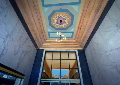 A well-lit, grand hallway with marble walls and a striking, geometric-patterned ceiling. large windows provide a view of reflected trees, enhancing the architectural aesthetics.