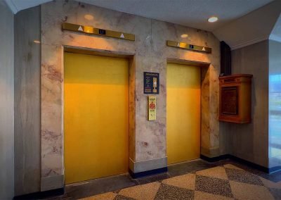 Two closed elevator doors with yellow panels and marble walls in an upscale building lobby, featuring geometric patterned flooring and a directory sign beside the doors.