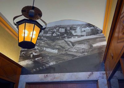 A vintage black-and-white panoramic cityscape mural on an arched wall beneath a light fixture with a warm glow, surrounded by elegant wood paneling in a hallway.