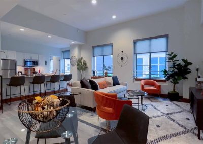 A spacious, modern living room with a neutral color palette featuring a sectional sofa, vibrant orange chairs, a kitchen in the background, and large windows that provide natural light. indoor plants add a touch of greenery.
