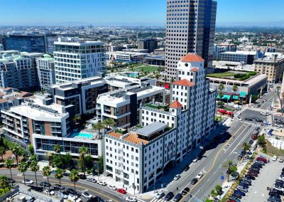 Aerial view of a bustling urban area with modern and traditional architecture, featuring a prominent building with a white facade and red-tiled roof, surrounded by roads and other structures.