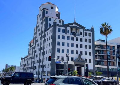 A modern, multi-story black and white building with a unique architectural design, featuring a tower-like structure with a dome, located in a sunny urban area with palm trees and traffic.