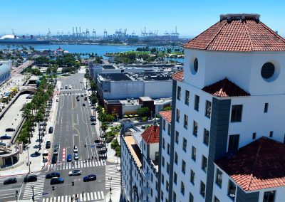 Aerial view of a cityscape with a prominent white building featuring red-tiled roofs. the background shows a street lined with palm trees and distant shipping cranes at a harbor.