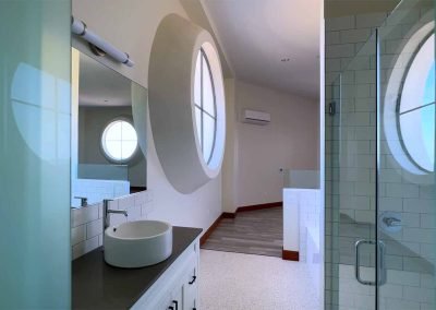 A bright, modern bathroom with a round mirror and a white vessel sink atop a white cabinet, green tiles, and a glass door reflecting the interior.