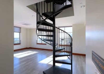 A spacious, well-lit room with hardwood floors, large windows, and a modern black spiral staircase in the center, leading to an upper level.