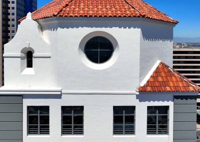 A white building with a red-tiled roof featuring a symmetrical design, a circular window, and a small bell tower, set against a backdrop of taller modern city buildings under a clear blue sky.