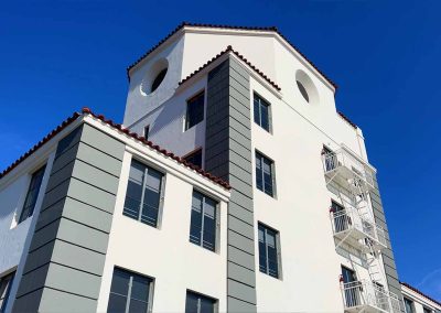 White three-story building with a tiled roof under a clear blue sky. the building has balconies and arched windows on the upper floors.