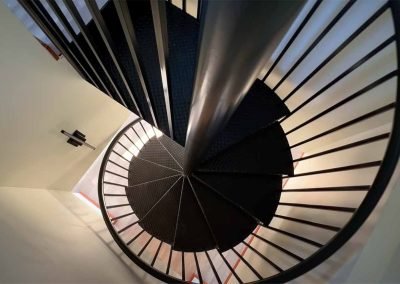 Spiral staircase viewed from above, featuring black steps and a central pole with a sleek metal handrail, contrasting with light-colored walls.
