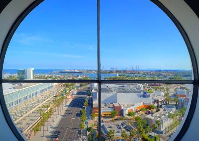 A scenic view through a circular window overlooking a cityscape with a busy street, buildings, and a distant port with cranes and ships under a clear blue sky.