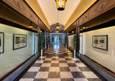 Elegant corridor with checkered floor tiles, adorned with framed historical photographs on either side, leading towards a brightly illuminated end under ornate hanging lamps.