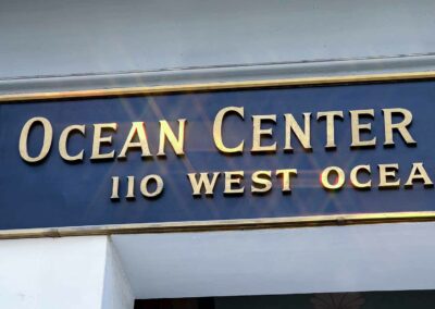 A close-up of a sign reading "ocean center building 110 west ocean" in raised golden letters against a dark blue background, with sunlight causing a lens flare on the upper part.